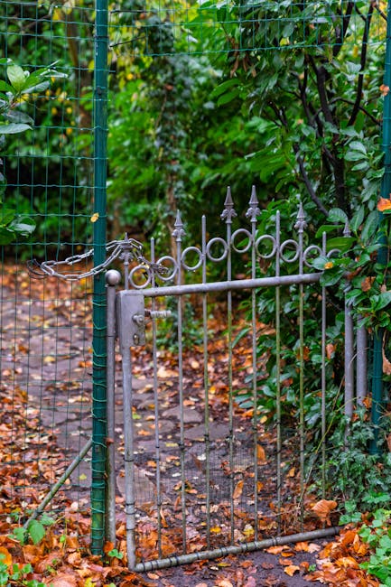 A small, decorative metal garden gate with ornate pointed finials on top, painted in a weathered gray finish, partially open and set within a green chain-link fence supported by a metal pole. The gate
