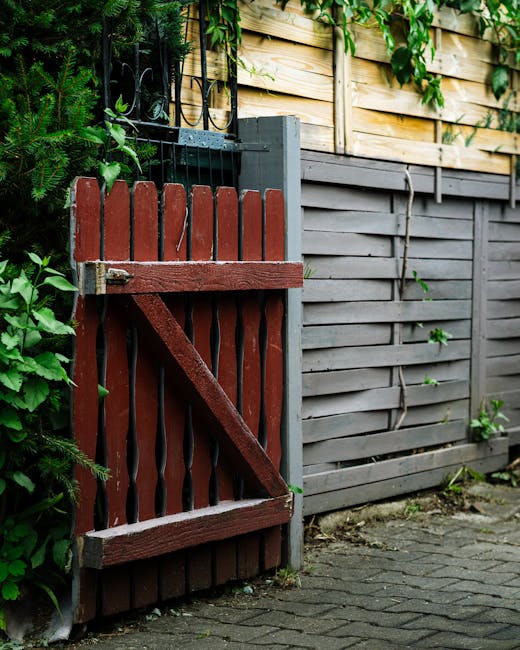 A small, decorative metal garden gate with ornate pointed finials on top, painted in a weathered gray finish, partially open and set within a green chain-link fence supported by a metal pole. The gate