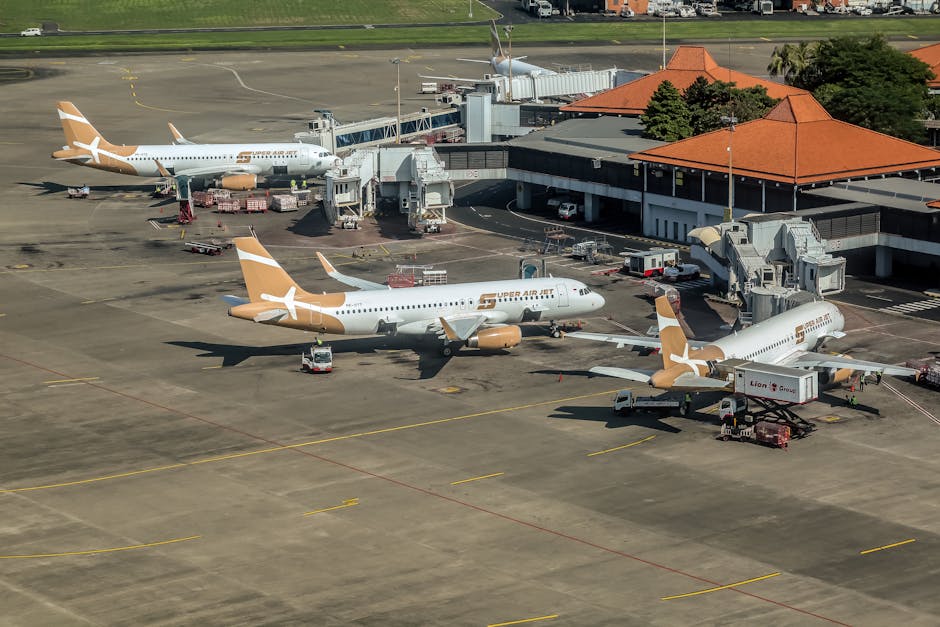 Aerial view of an airport tarmac showing three white and tan aircraft parked at the gates, with jet bridges connecting them to a terminal building with a red-tiled roof. The foreground features wet co